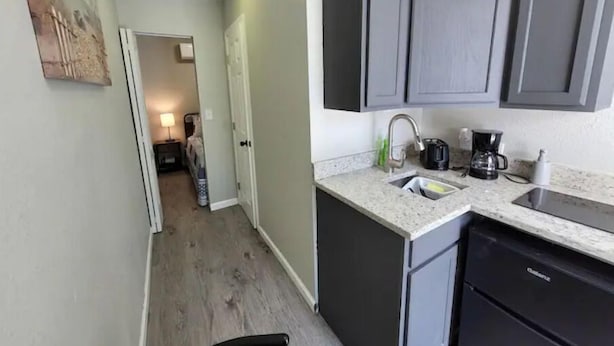 Una cocina compacta con armarios oscuros, una encimera y un lavavajillas. Un pasillo conduce a un dormitorio con una cama y una mesita de noche.