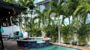 Una piscina con idromassaggio, circondata da palme e un patio con sedie e ombrelloni.