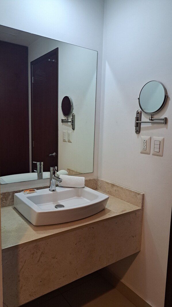 Un baño con un lavamanos blanco, un espejo grande, un espejo redondo y una toalla colgada en la pared.