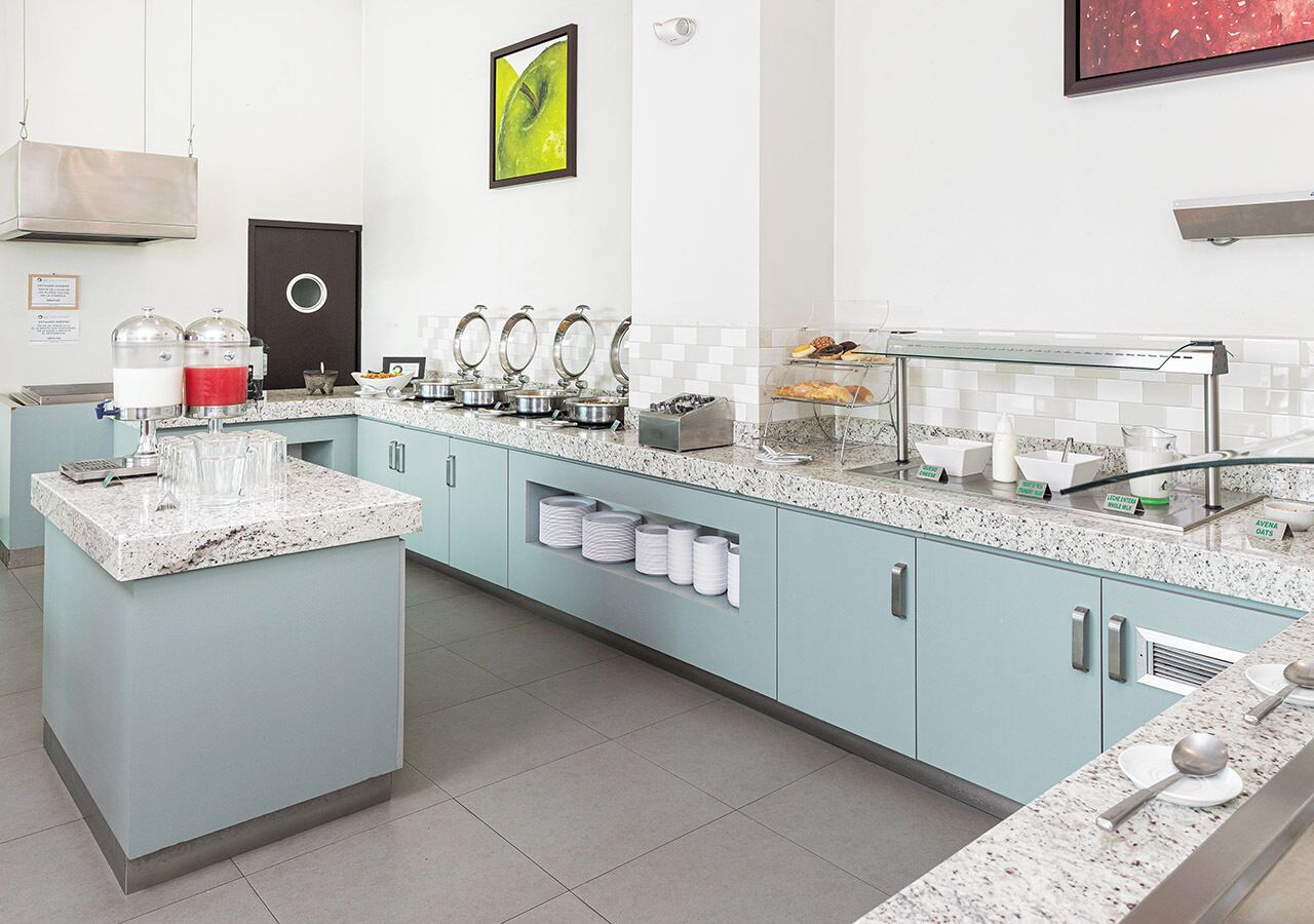 Una cocina moderna con alacenas azul claro, encimeras de granito y electrodomésticos de acero inoxidable. Hay varias estaciones de servicio con recipientes de comida y bebida, además de una máquina de café.