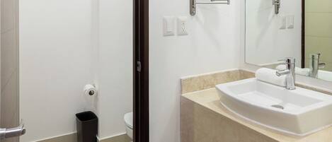 A bathroom with a sink, mirror, toilet, and trash bin.
