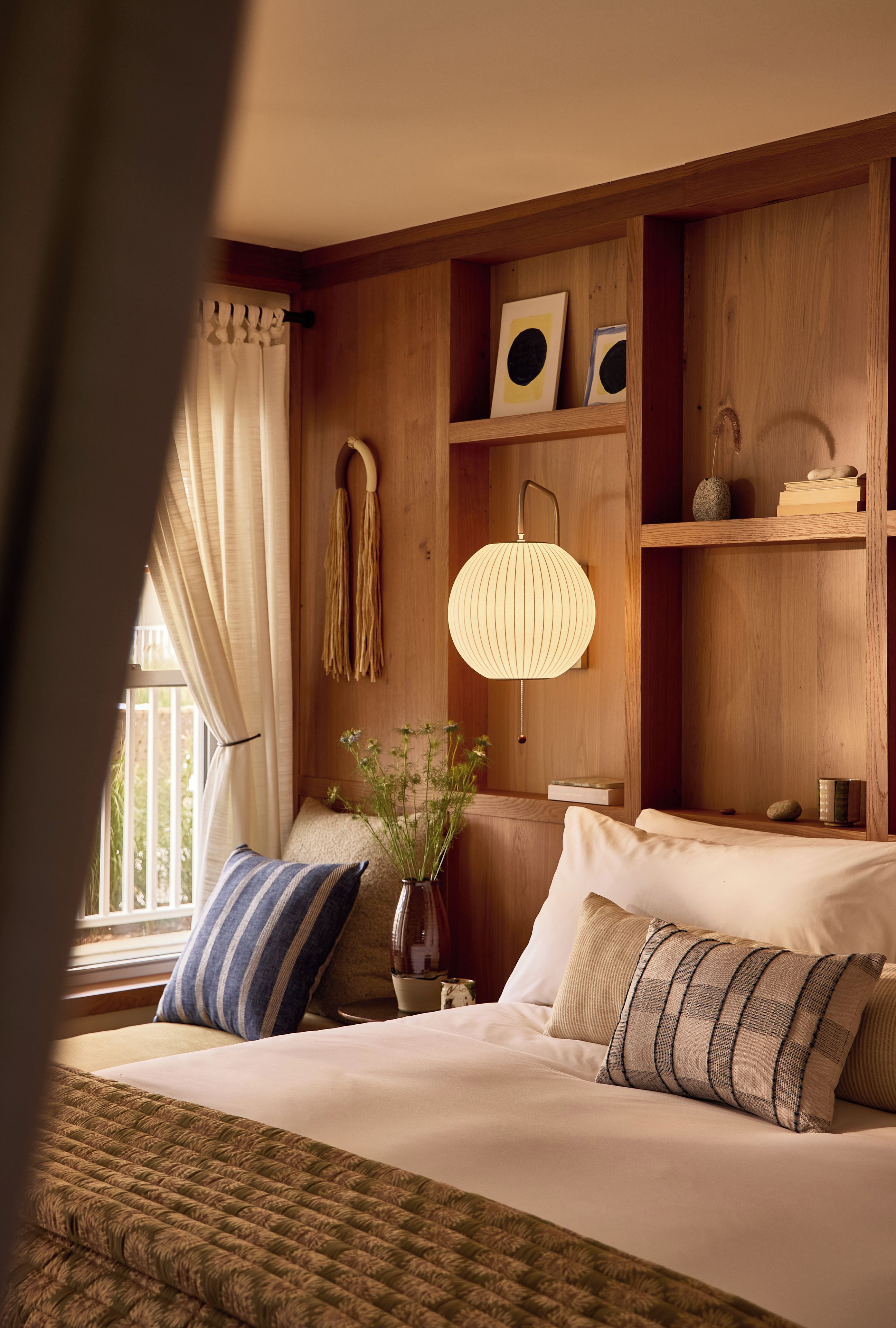Habitación de hotel con cama, almohadas, lámpara colgante, un florero con plantas y estanterías de madera con libros y objetos decorativos.
