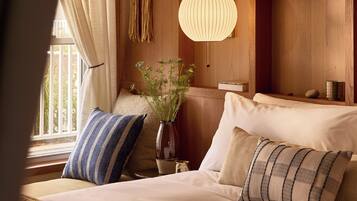 Habitación de hotel con cama, almohadas, lámpara colgante, un florero con plantas y estanterías de madera con libros y objetos decorativos.