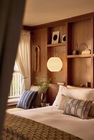 Habitación de hotel con cama, almohadas, lámpara colgante, un florero con plantas y estanterías de madera con libros y objetos decorativos.