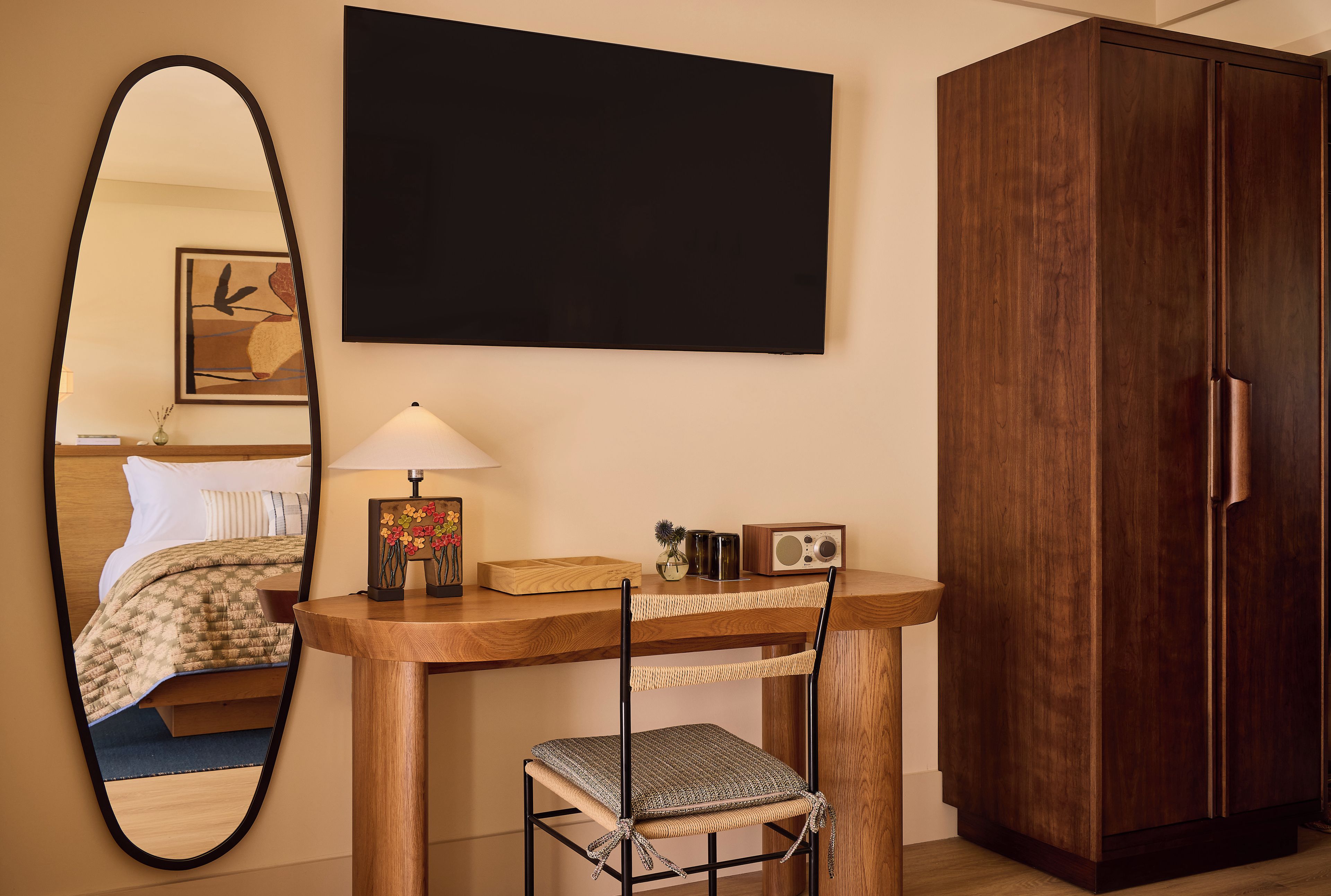 A hotel room with a bed, a desk with a lamp, a chair, a mirror, and a wardrobe.