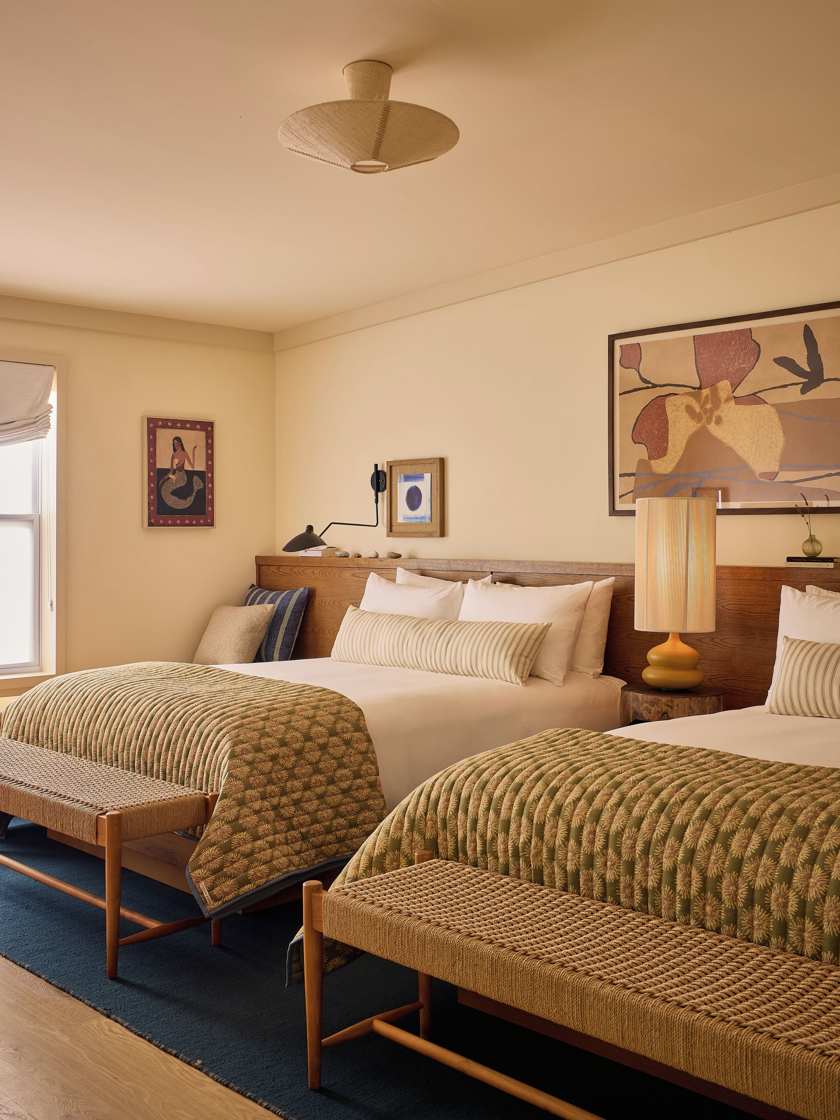A hotel room with two beds, a large window, a ceiling fan, and a painting on the wall.