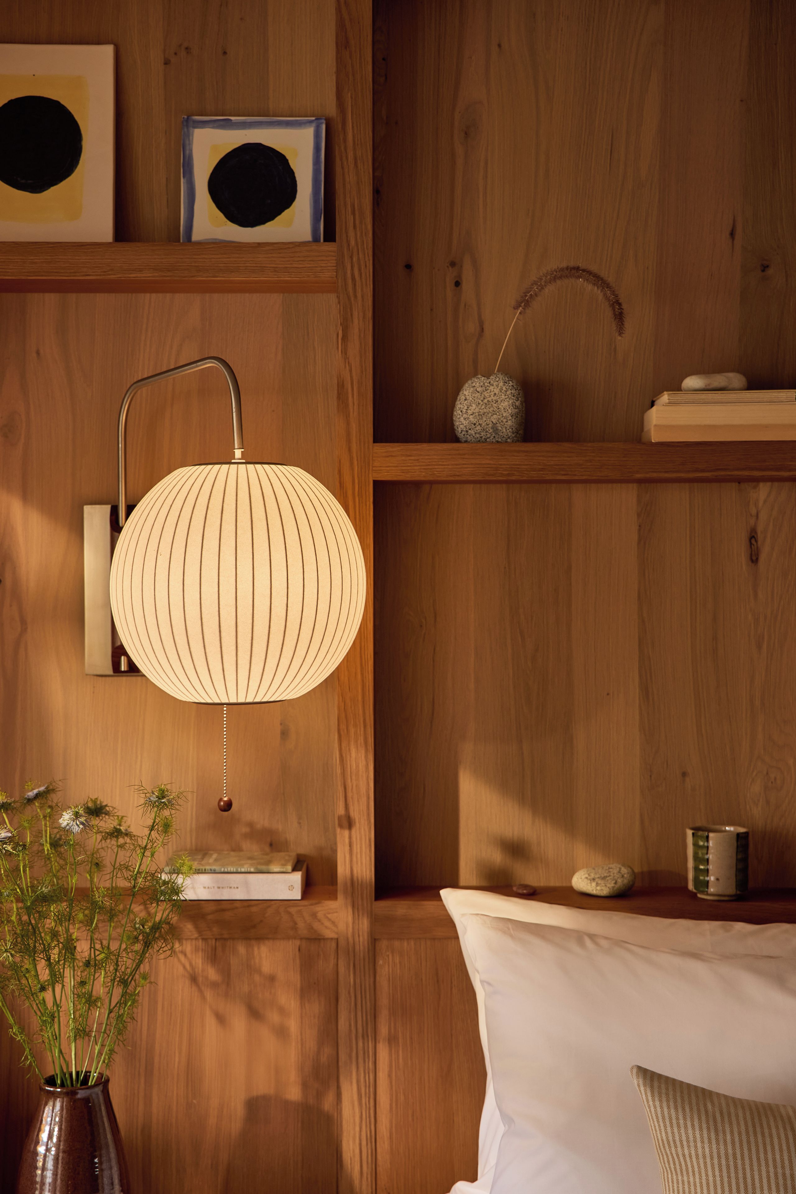 A wooden wall with a wall-mounted lamp, a shelf with decorative items, and a bedside table with a pillow.