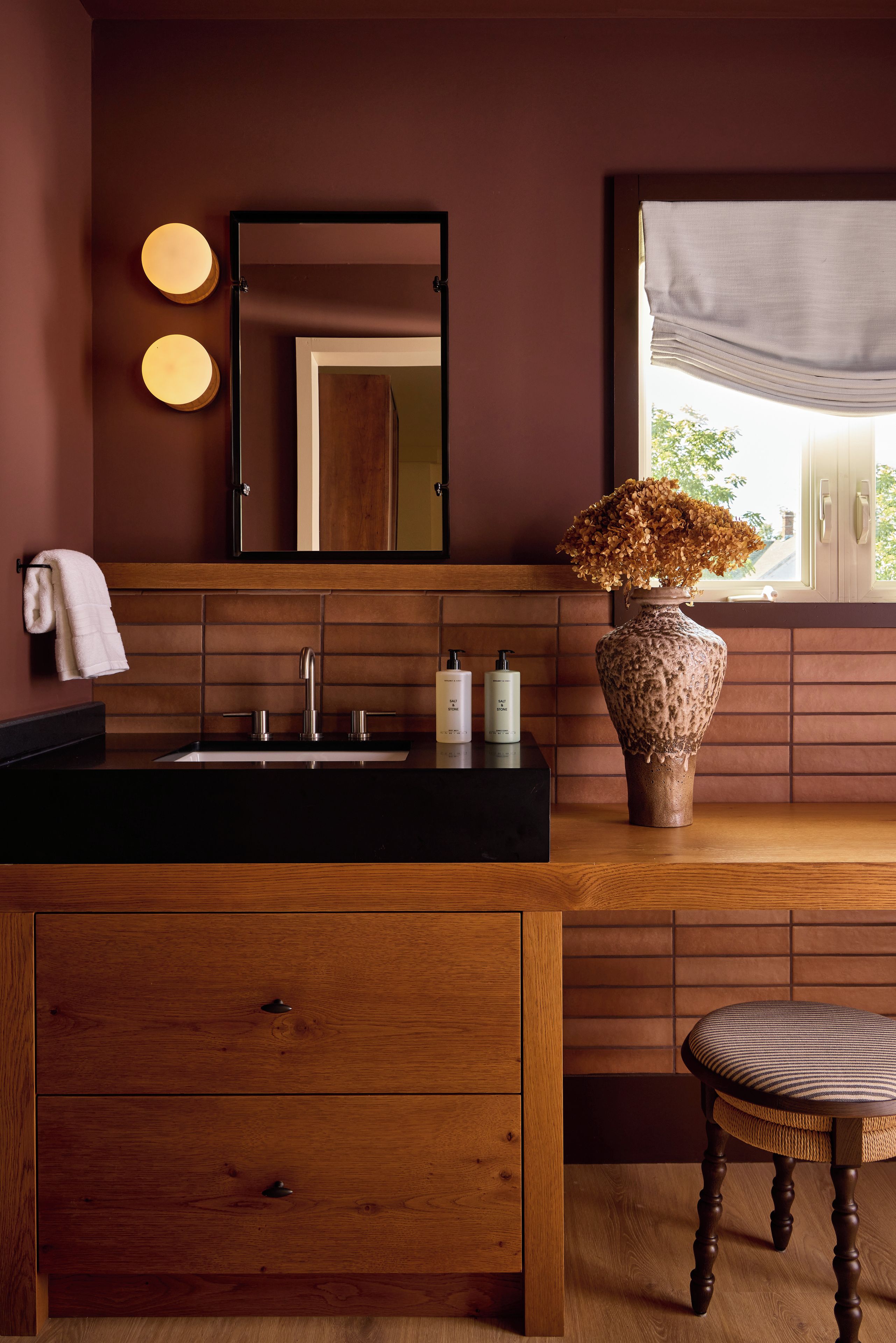 Une salle de bain avec un meuble-lavabo en bois, un miroir, un lavabo avec un plan de travail noir, deux bouteilles de savon, un vase avec des fleurs séchées et une fenêtre donnant sur un paysage verdoyant.