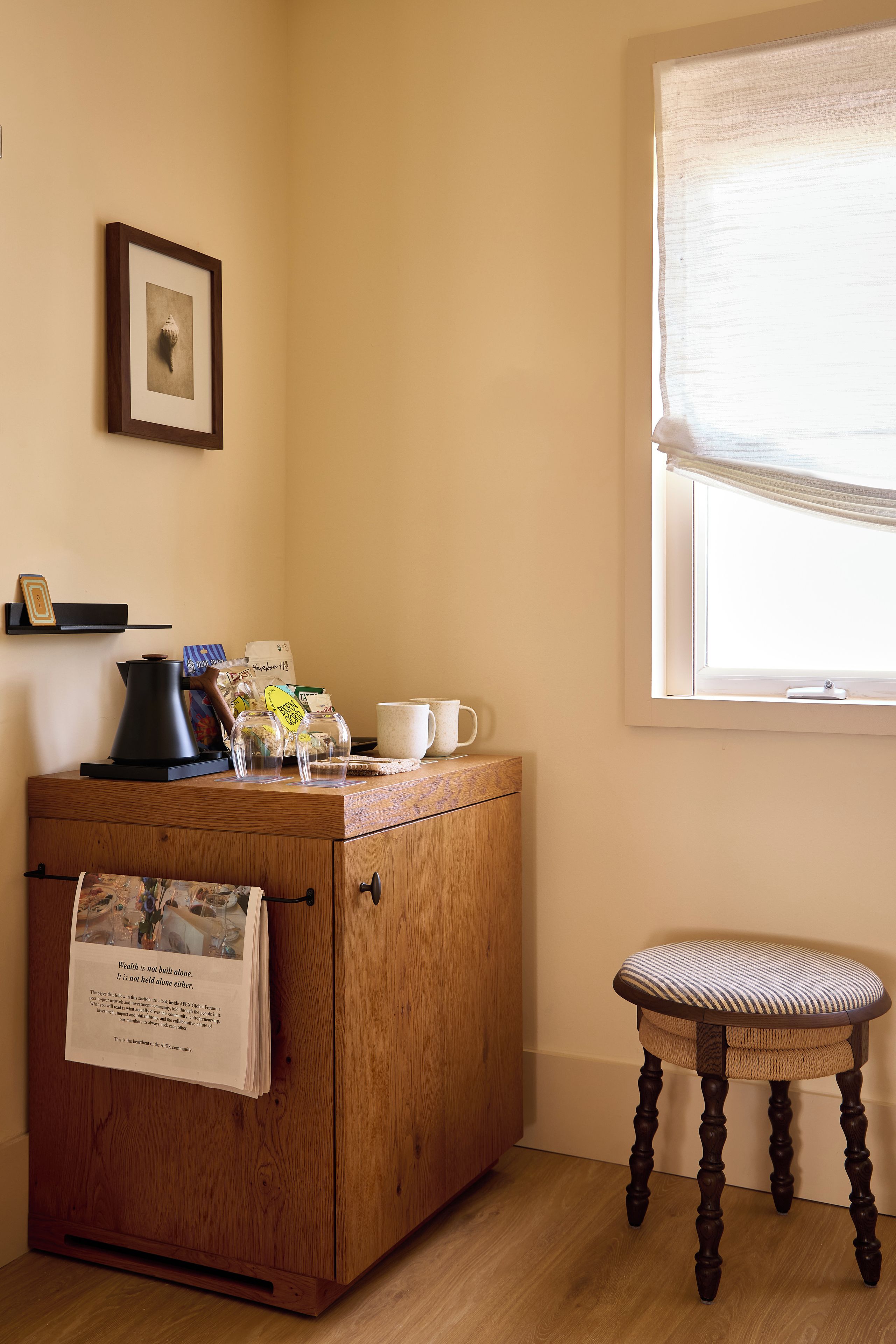 A wooden cabinet with a newspaper, a coffee maker, and a mug on top. There is a small stool and a window with blinds.