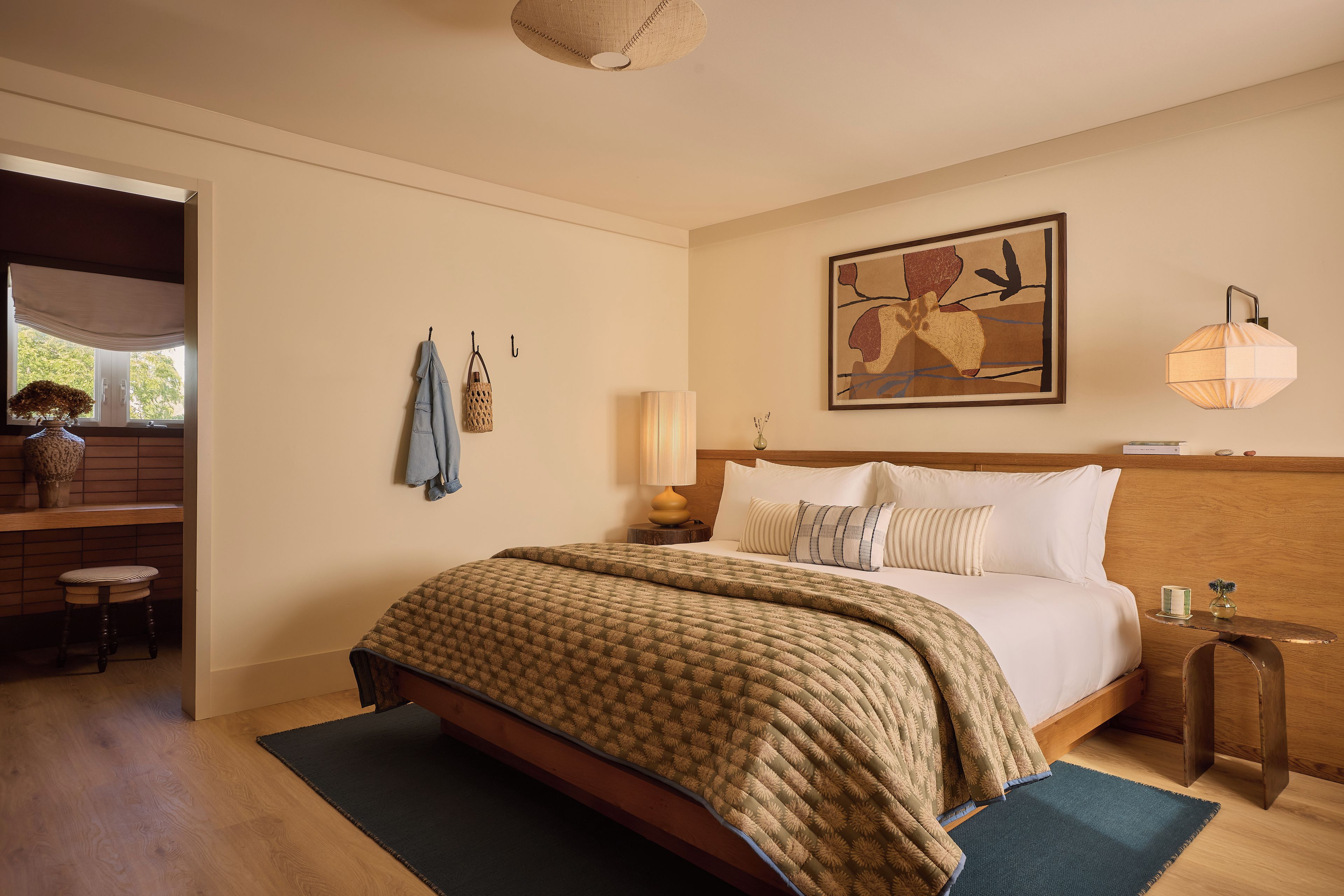 A bedroom with a bed, bedside table, lamp, artwork, and a view of the outdoors.