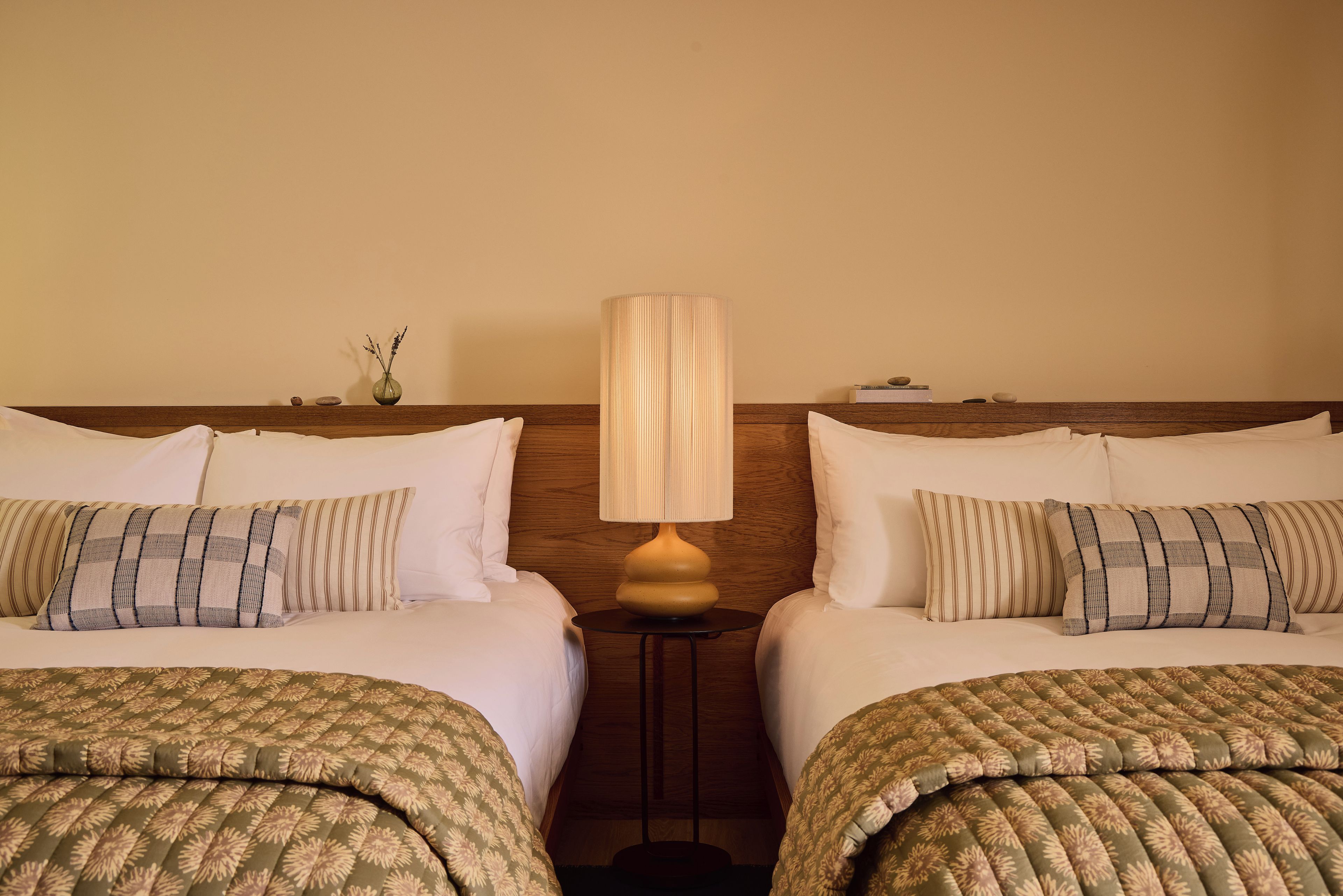A hotel room with two beds, a bedside table with a lamp, and a vase with flowers.