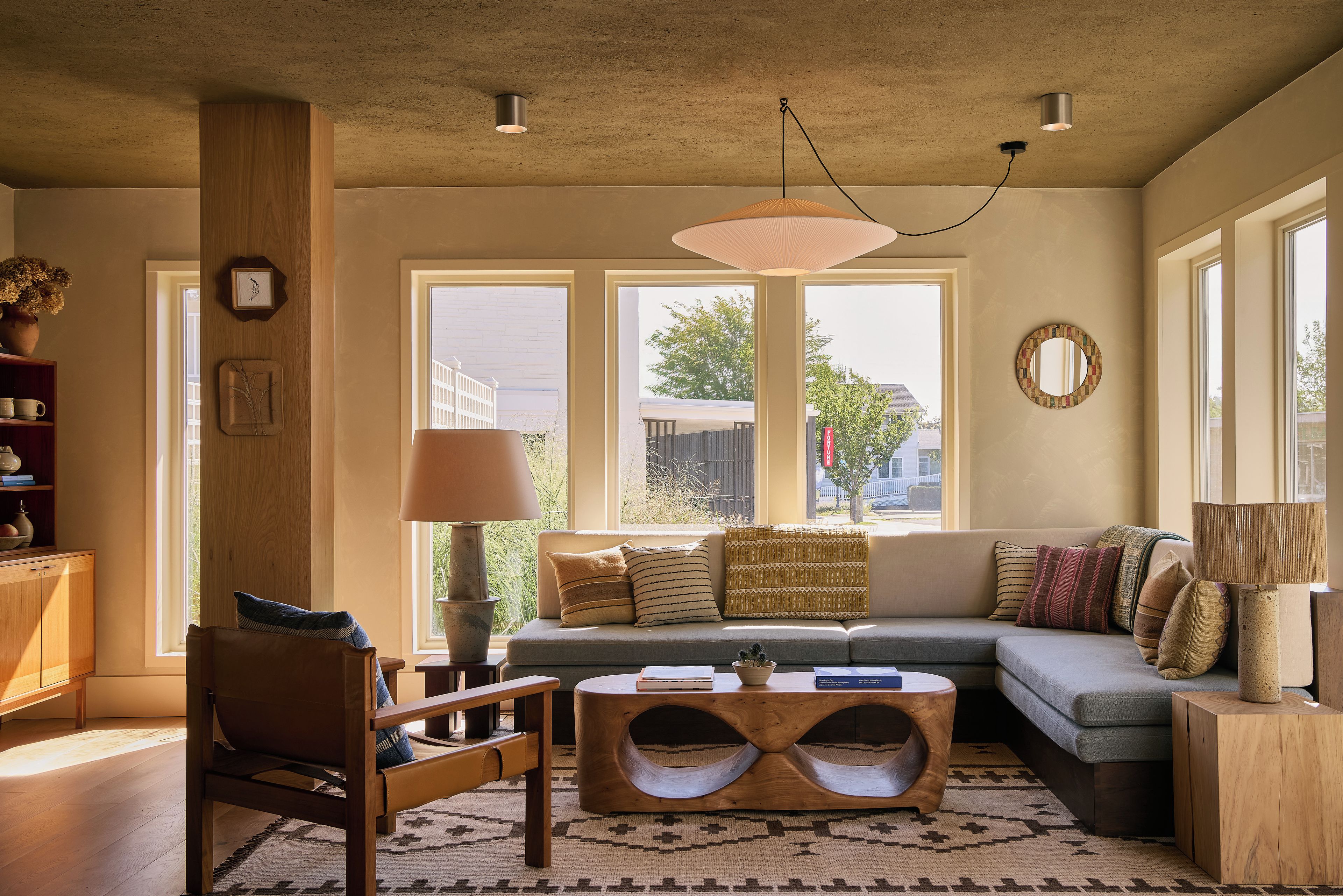 A living room with a sofa, a wooden coffee table, a chair, and a lamp. There are windows with a view of the outside.