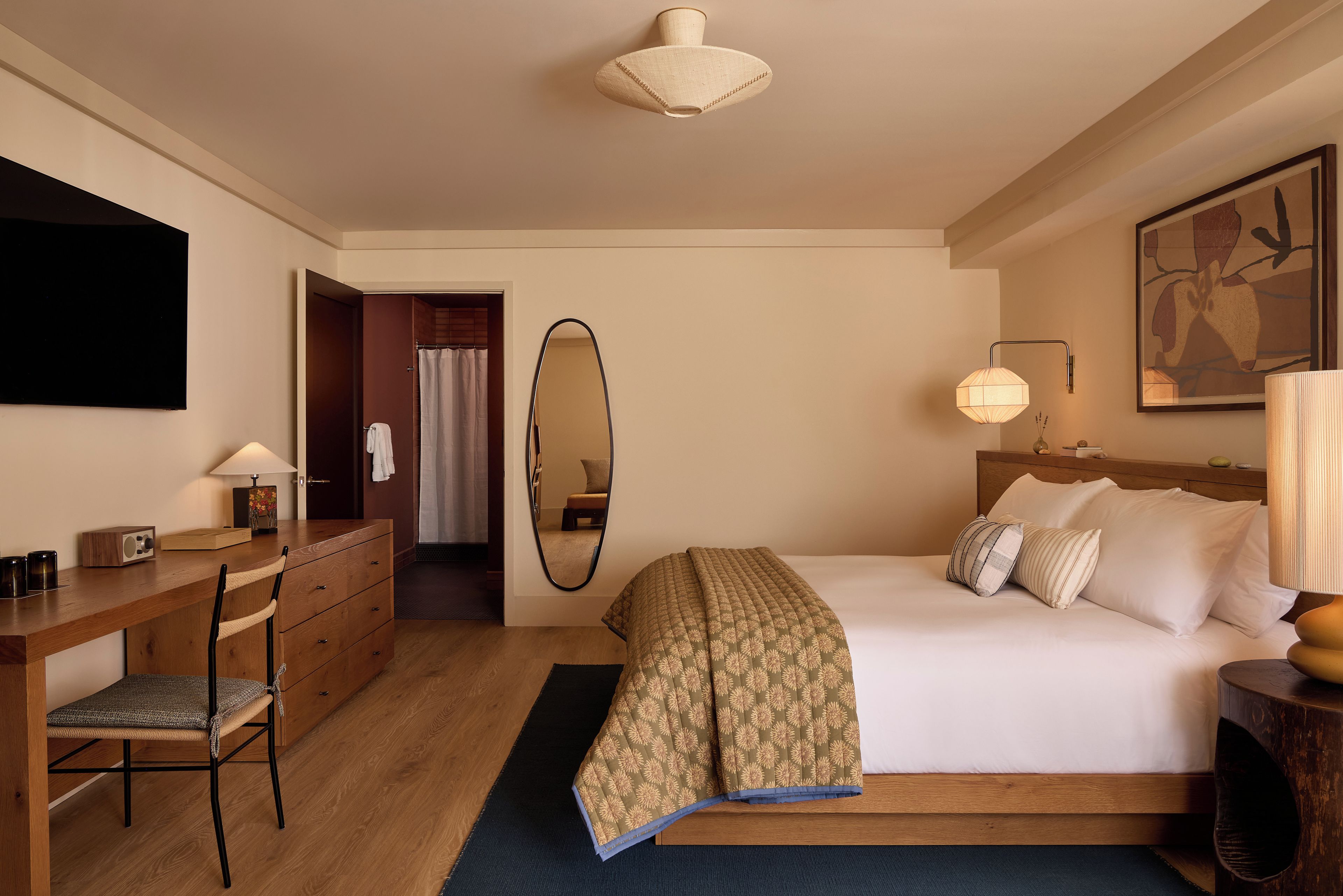 A hotel room with a bed, bedside table, lamp, chair, desk, and a mirror.