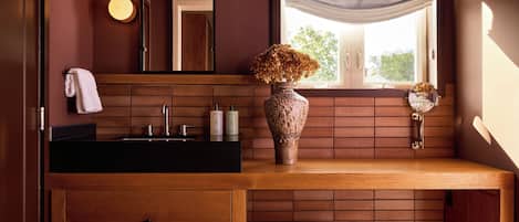 Une salle de bain avec un meuble-lavabo en bois, un miroir, un vase contenant des fleurs séchées et une fenêtre donnant sur des arbres.