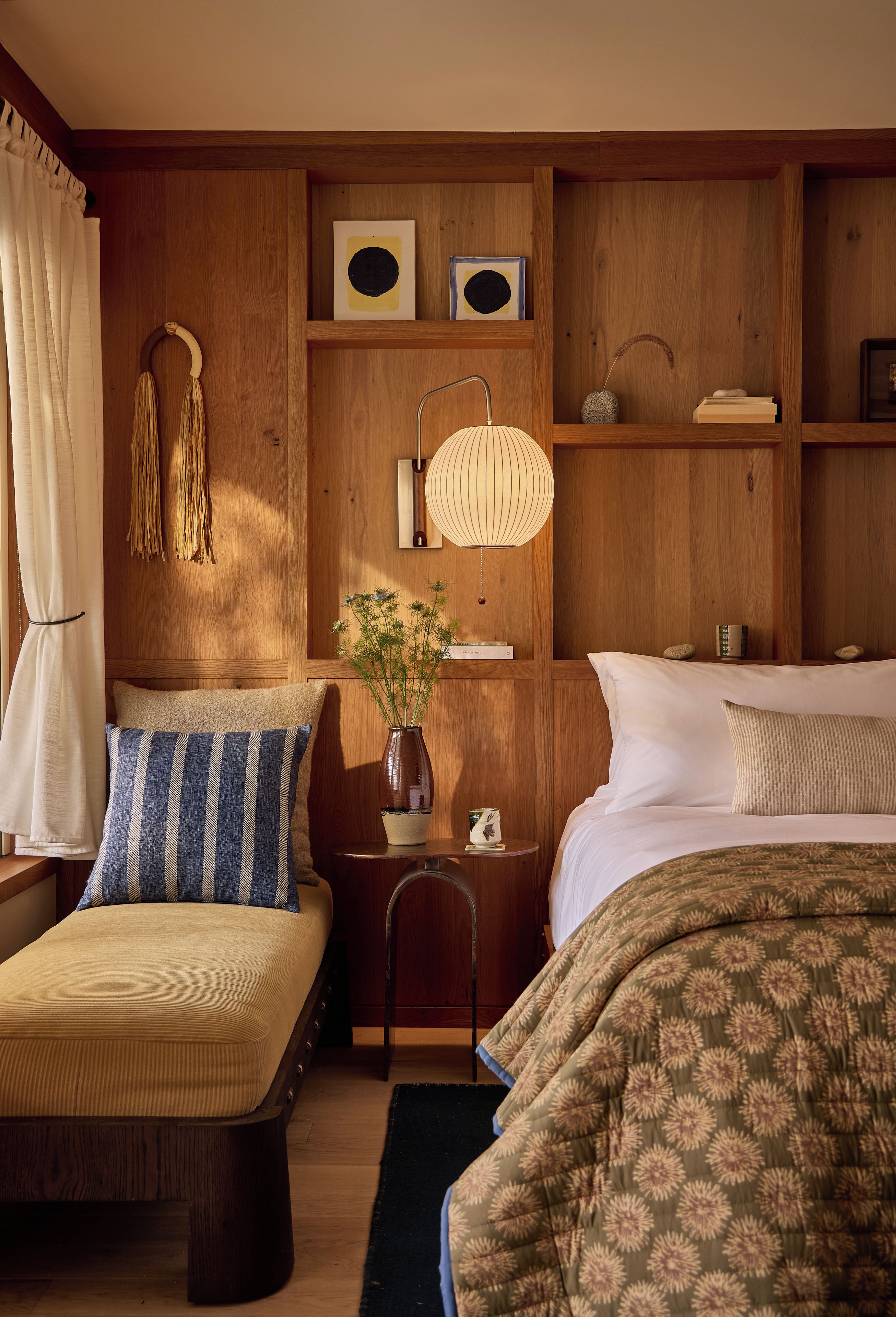 Un dormitorio acogedor con cabecera de madera, una mesita de noche con lámpara, un jarrón con flores y una cama con colcha estampada.