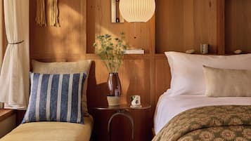 Un dormitorio acogedor con cabecera de madera, una mesita de noche con lámpara, un jarrón con flores y una cama con colcha estampada.