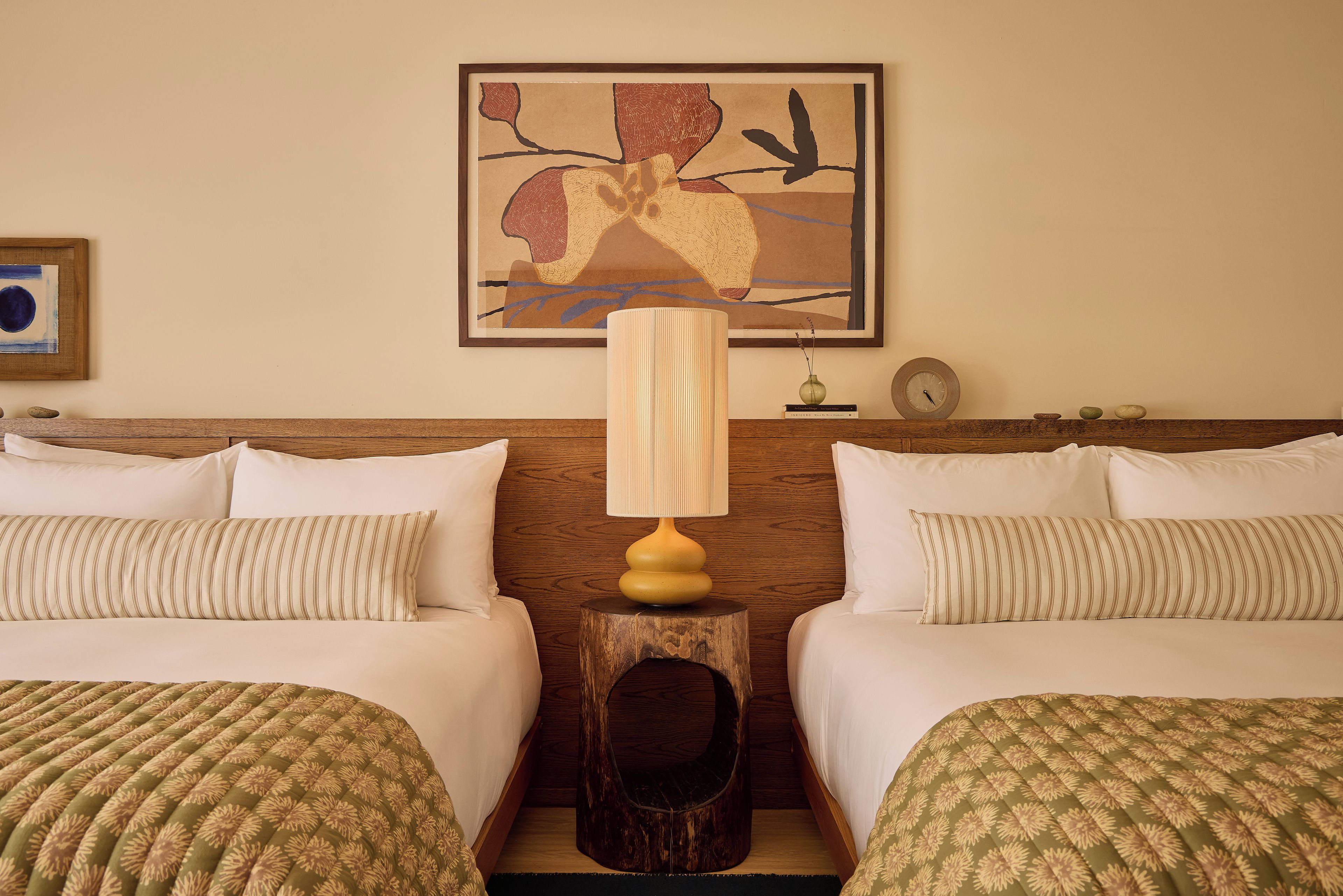 A hotel room with two beds, a wooden headboard, a lamp, a clock, and a framed artwork on the wall.