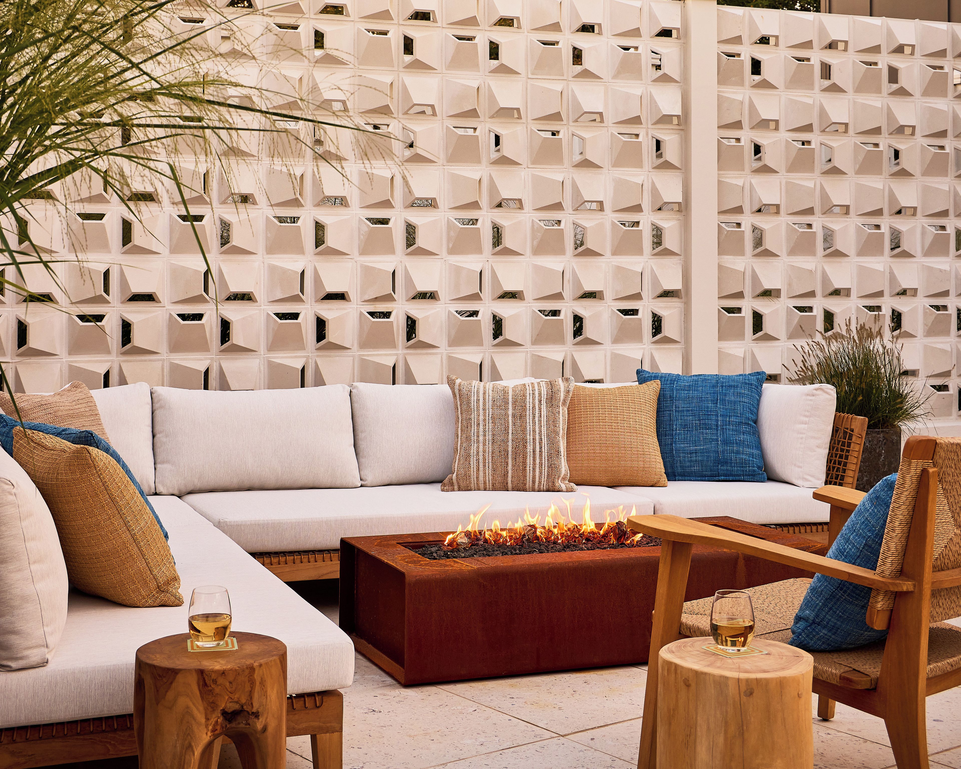 A modern outdoor seating area with a sofa, cushions, a wooden coffee table with a fire feature, and a decorative wall with geometric patterns.