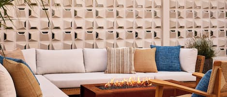 Un espace de détente extérieur moderne, comprenant un canapé, des coussins, une table basse en bois dotée d’un foyer, et un mur décoratif orné de motifs géométriques.