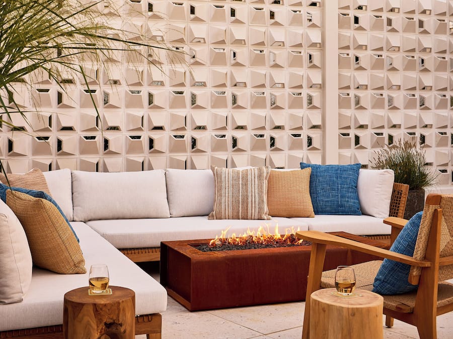 A modern outdoor seating area with a sofa, cushions, a wooden coffee table with a fire feature, and a decorative wall with geometric patterns.