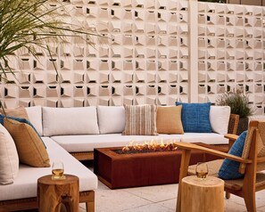 Un área de estar al aire libre y moderna, con un sofá, cojines, una mesa de centro de madera con chimenea y una pared decorativa con patrones geométricos.