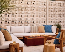 A modern outdoor seating area with a sofa, cushions, a wooden coffee table with a fire feature, and a decorative wall with geometric patterns.