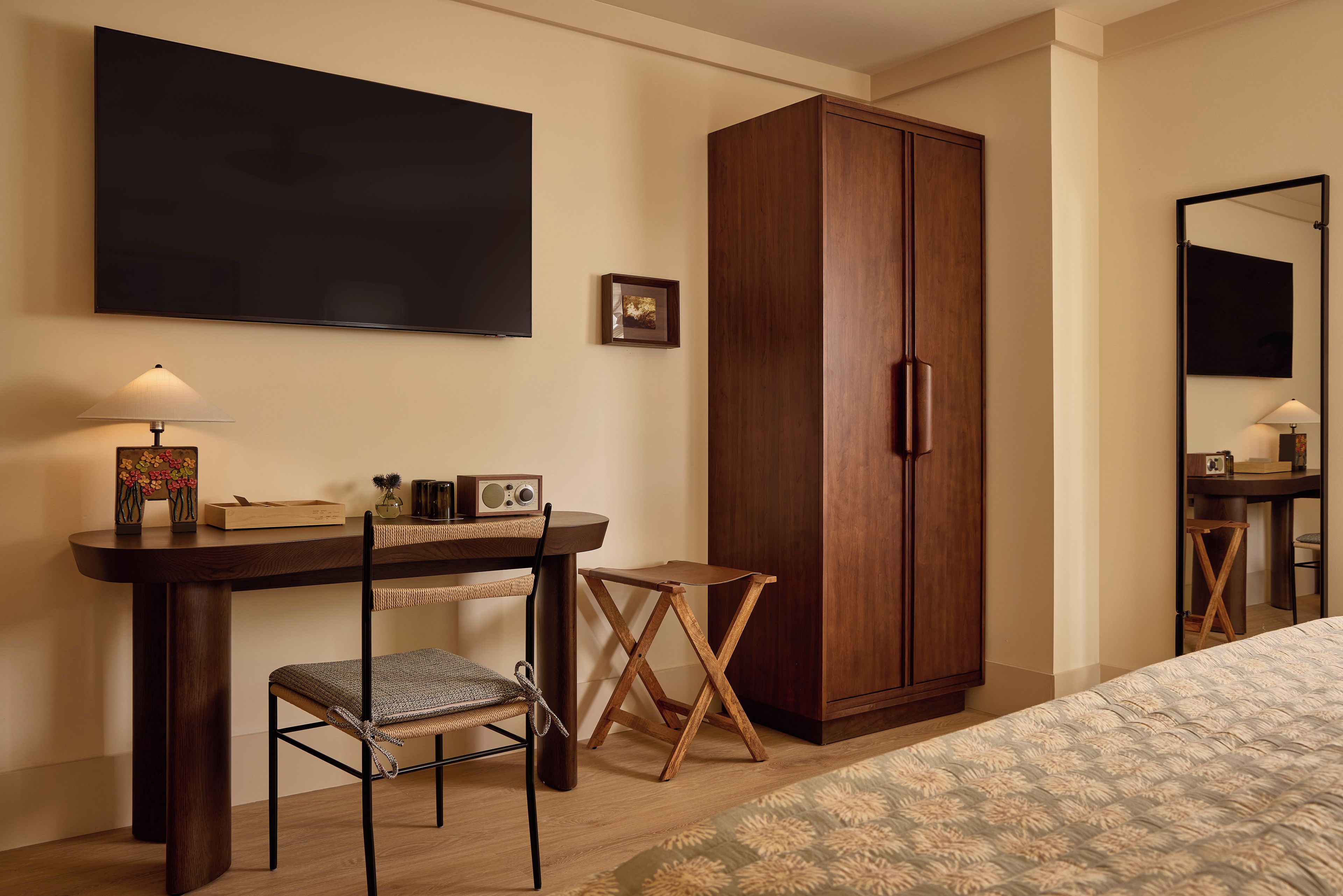 A hotel room with a bed, a desk with a chair, a wardrobe, a television, and a small table with a lamp.