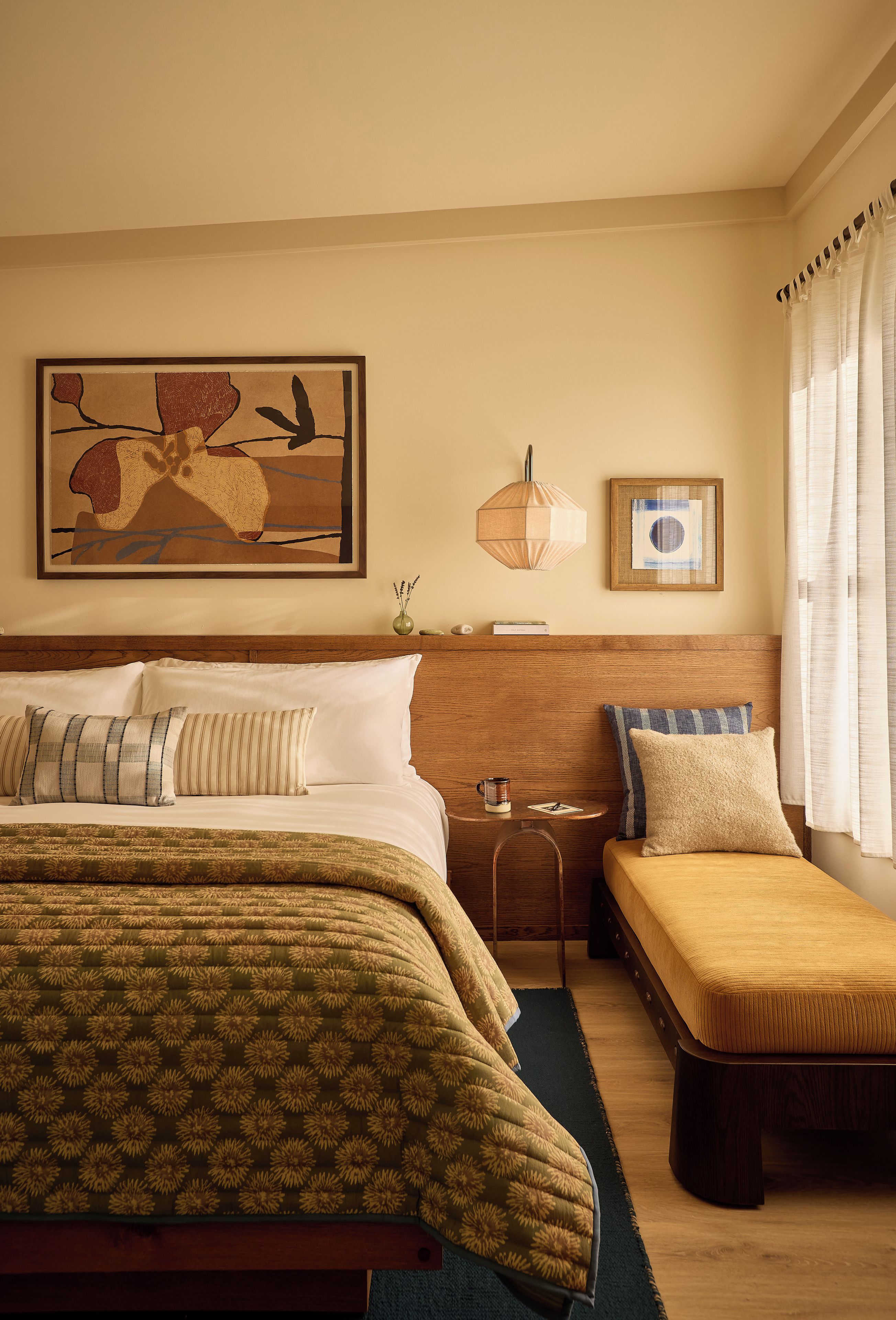 A hotel room with a large bed, a smaller sofa, a bedside table with a lamp, and a framed artwork on the wall.
