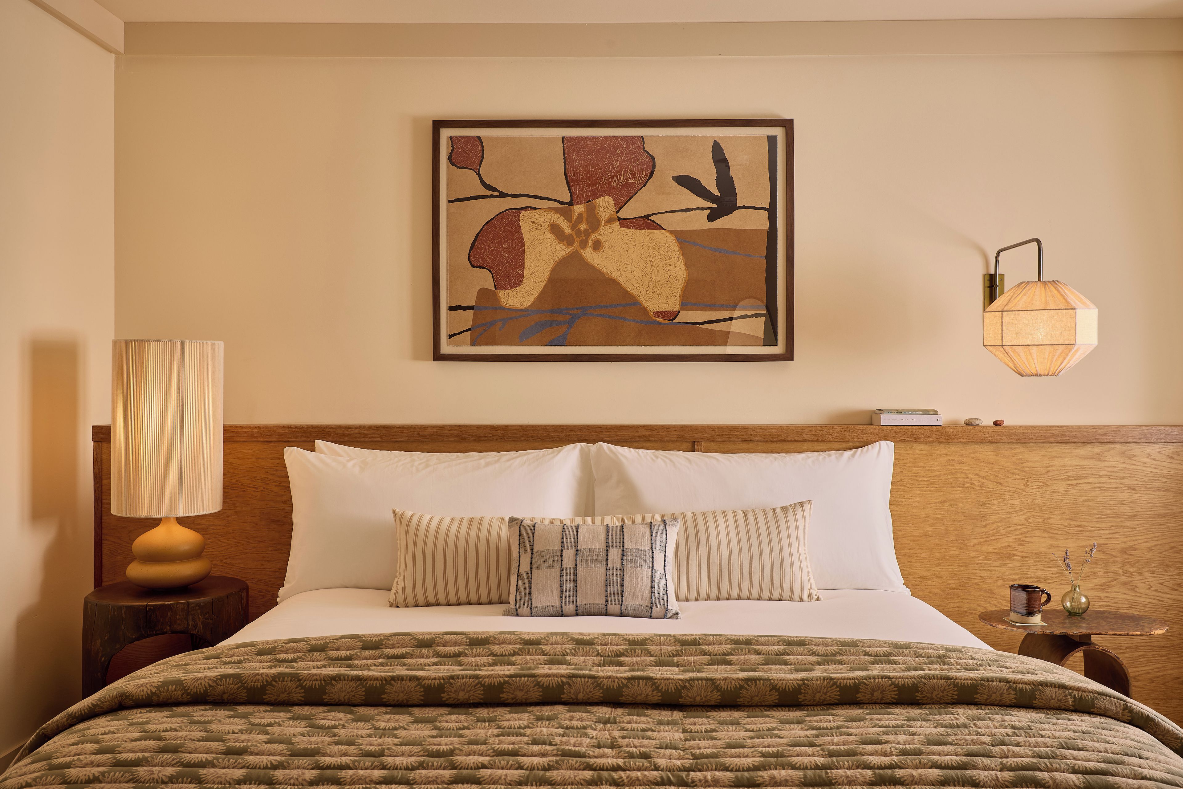 Habitación de hotel con cama, almohadas, lámpara, una obra de arte en la pared y una mesita de noche.