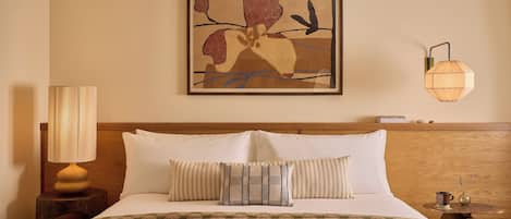 A hotel room with a bed, pillows, a lamp, a wall art piece, and a nightstand.