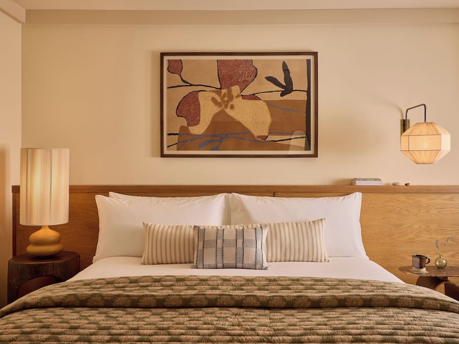 A hotel room with a bed, pillows, a lamp, a wall art piece, and a nightstand.