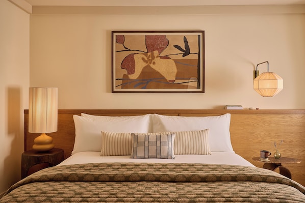 Habitación de hotel con cama, almohadas, lámpara, una obra de arte en la pared y una mesita de noche.