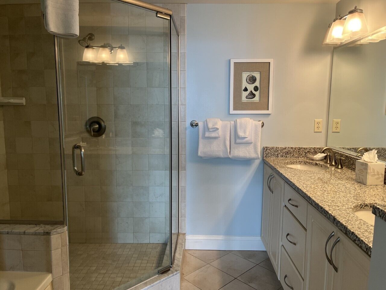 A bathroom with a glass shower enclosure, a sink with a granite countertop, and a framed picture on the wall.