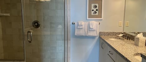 A bathroom with a glass shower enclosure, a sink with a granite countertop, and a framed picture on the wall.