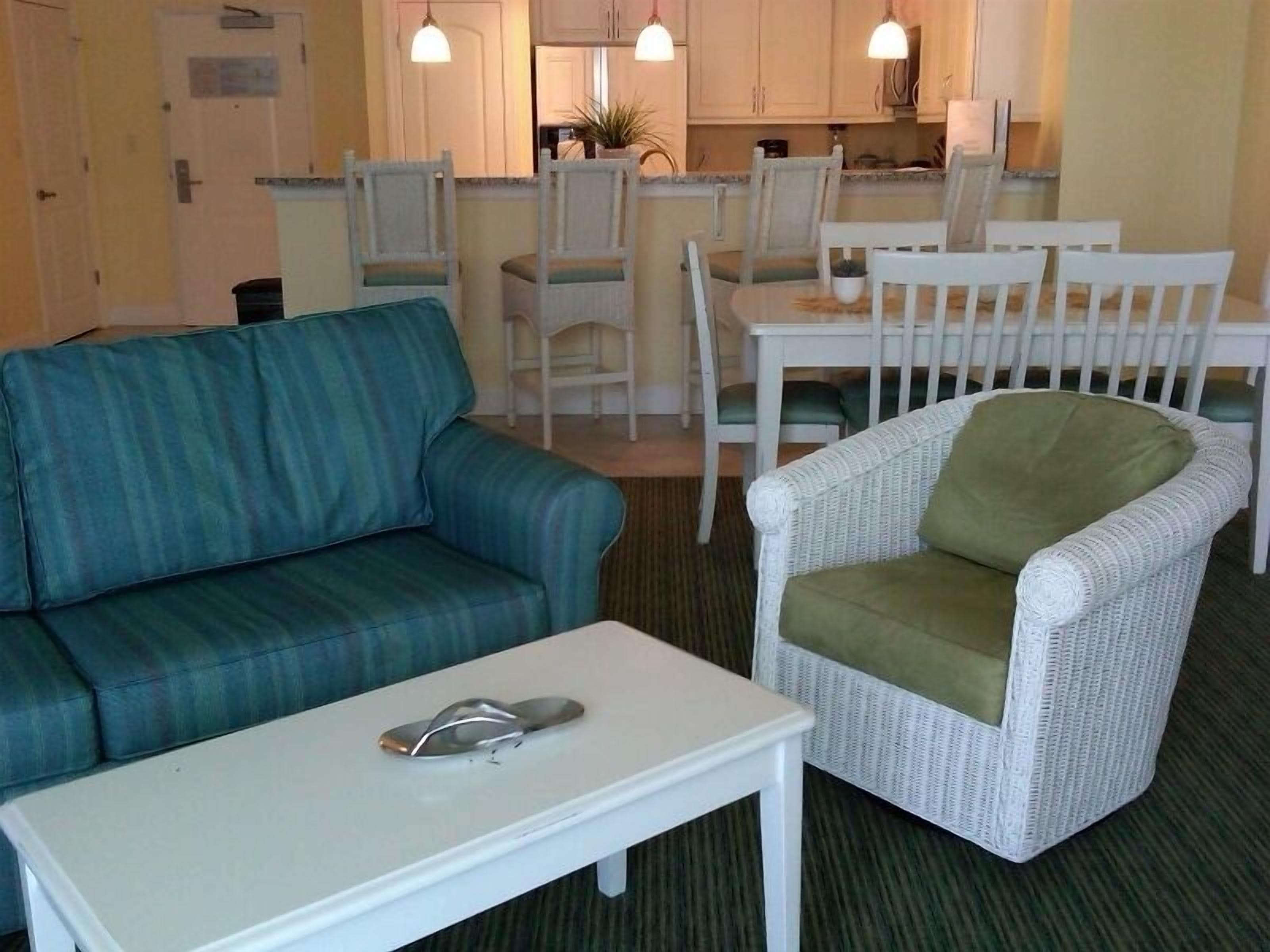 A living room with a blue sofa, a white coffee table, and a wicker armchair with a green cushion. There is a kitchen in the background with white cabinets and pendant lights.