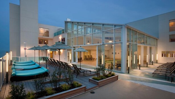 A modern building with a glass facade, outdoor seating area with umbrellas, and a pool. - Miramar Condominiums (Ocean City)