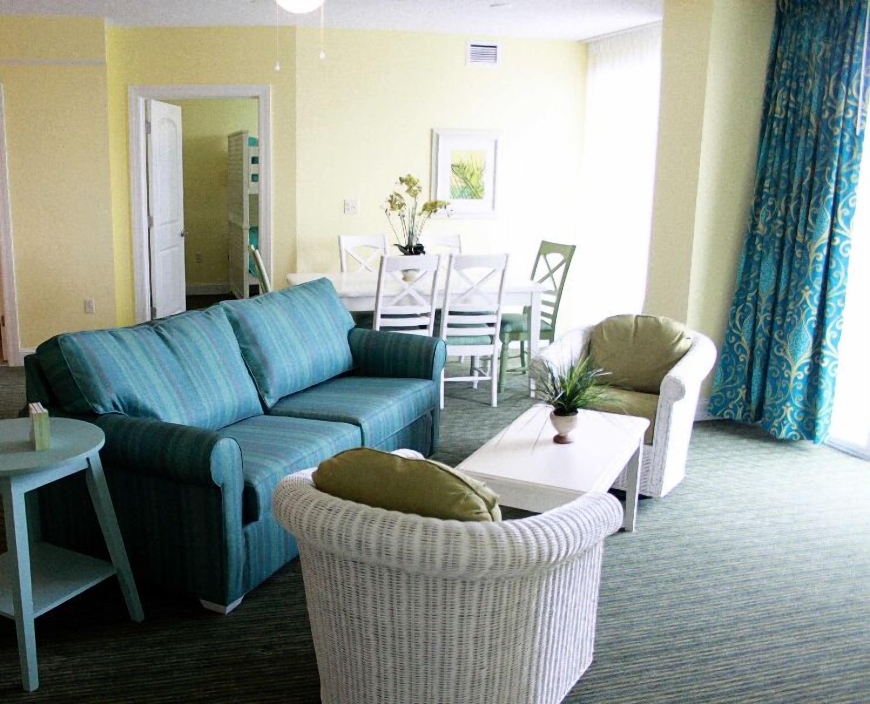 A room with a blue sofa, a white wicker chair, a small table with a plant, and a dining area with white chairs and a table.