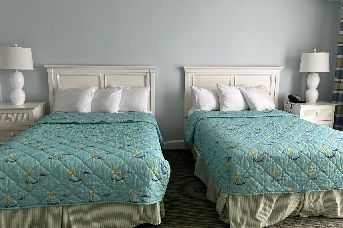 Habitación de hotel con dos camas, cada una con cabecero blanco y colcha acolchada color verde azulado. Hay mesitas de noche blancas con lámparas, alfombra a rayas y cortina a rayas en la ventana.