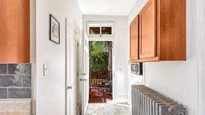 Un pasillo estrecho con suelo de baldosas, un radiador, armarios de madera y una puerta que da a un patio con mobiliario exterior.