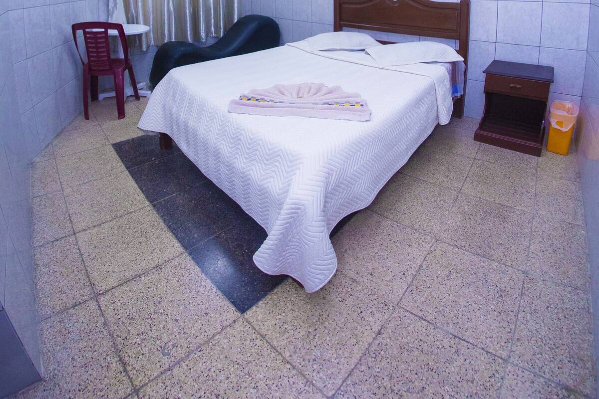 Ein Schlafzimmer mit Bett, Stuhl, Nachttisch, Mülleimer und Deckenventilator.