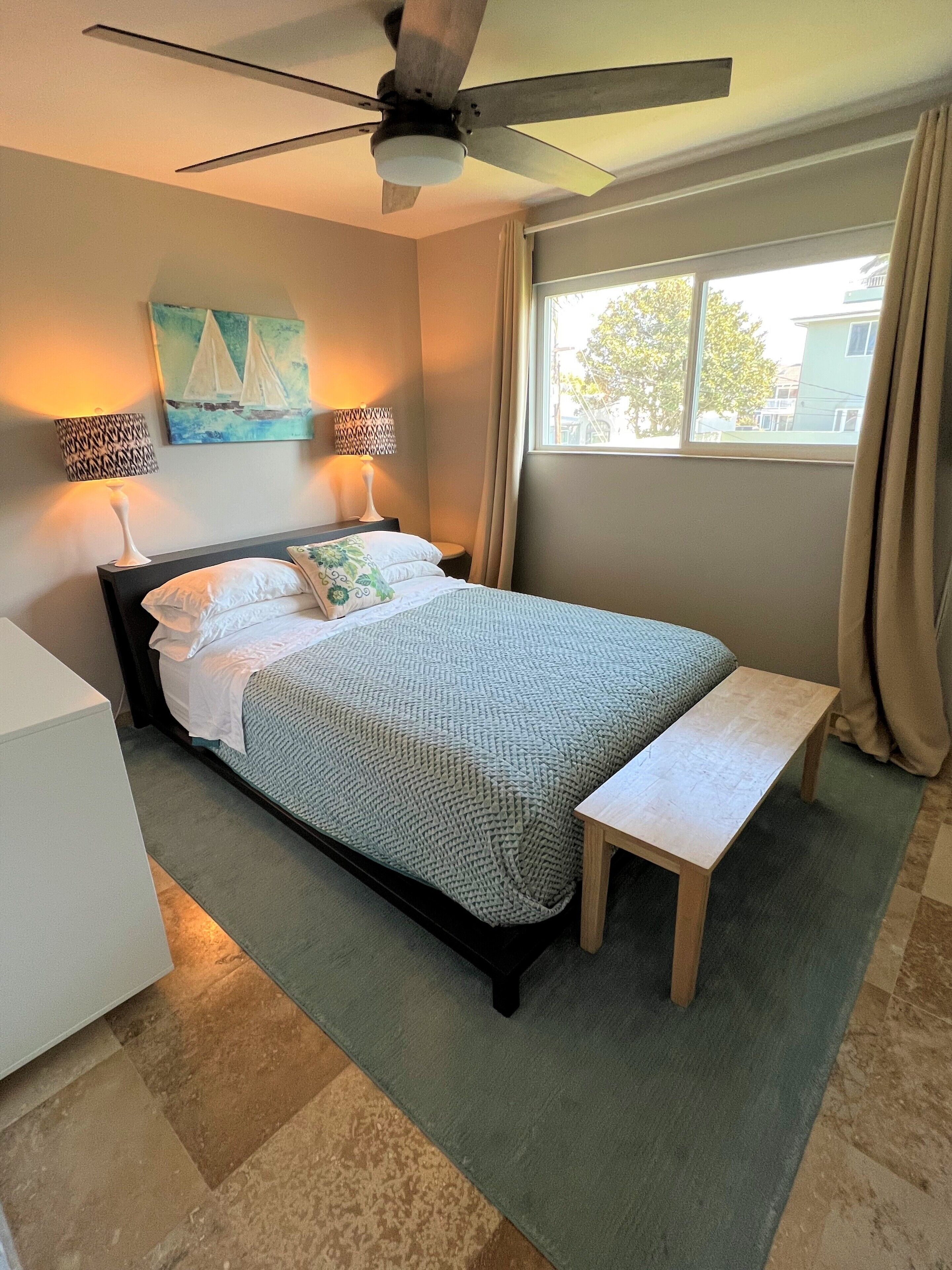 A neatly arranged bedroom with a bed, bedside lamps, a ceiling fan, a window with curtains, and a painting on the wall.