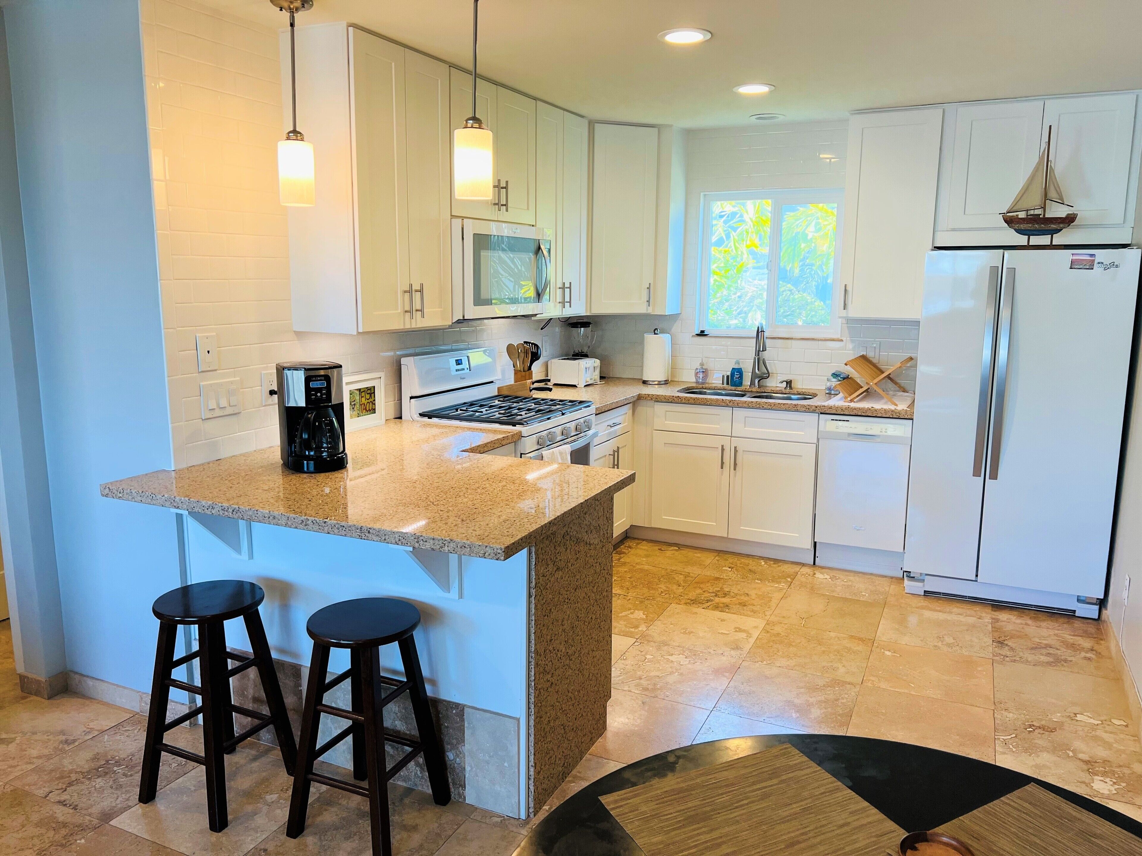 A modern kitchen with white cabinets, a granite countertop, a built-in oven, a sink, and a refrigerator. There are two bar stools and pendant lights above the counter.