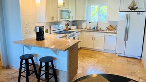 A modern kitchen with white cabinets, a granite countertop, a built-in oven, a sink, and a refrigerator. There are two bar stools and pendant lights above the counter.
