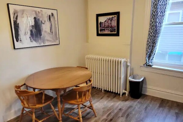 A room with a wooden table and chairs, a radiator, a framed picture, and a window with a curtain.