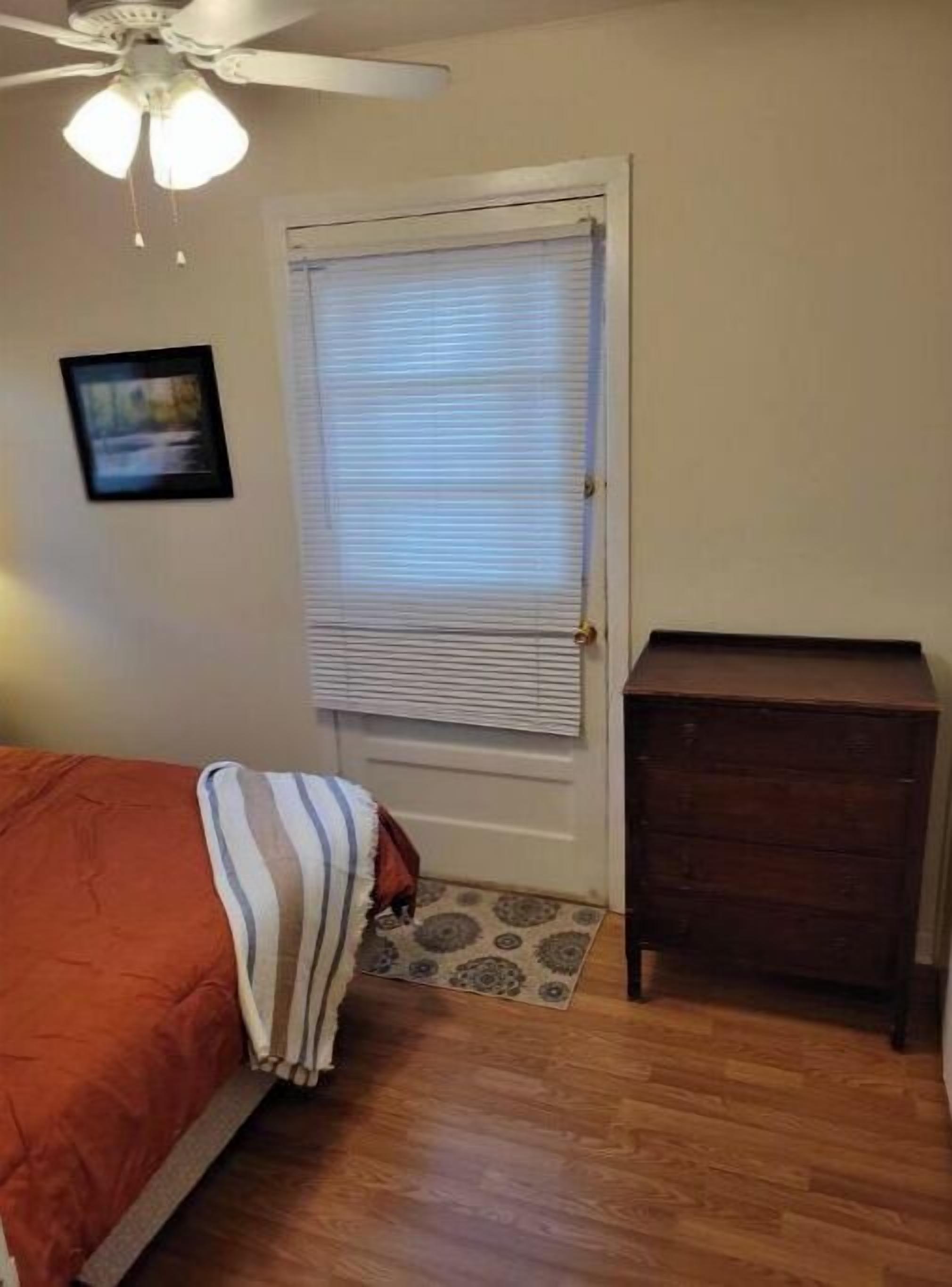 Une chambre à coucher comprenant un lit, un ventilateur de plafond, une fenêtre avec des stores, une petite commode en bois et un tapis.
