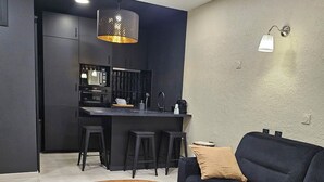 Un saló modern amb una taula de centre rodona de fusta, un sofà negre i una zona de cuina amb armaris foscos i un tamboret.
