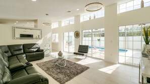 Ein geräumiges Wohnzimmer mit Ecksofa, Glastisch und Blick auf einen Swimmingpool durch große Fenster.