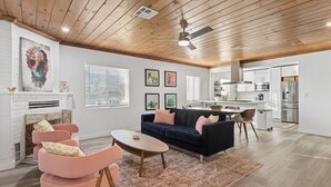 A spacious living room with a dark blue sofa, pink armchairs, and a wooden coffee table. A fireplace with a white brick wall and a ceiling fan are visible. The kitchen area is in the background with white cabinets and stainless steel appliances. - Freshly Renovated Midtown Phoenix Retreat with Pool (Phoenix)