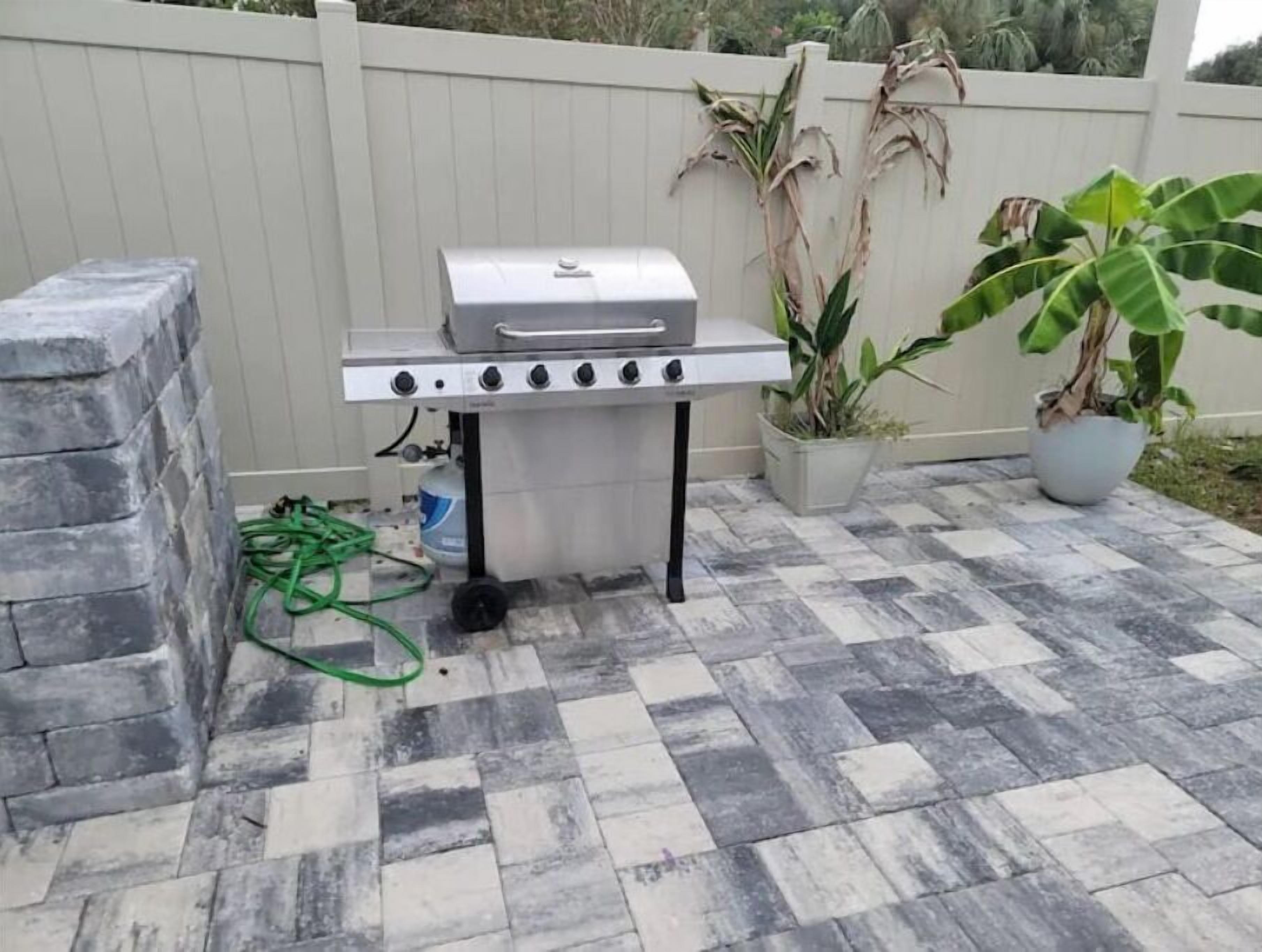 A portable barbecue grill with four burners, a water supply hose, and a gas canister on a tiled patio.