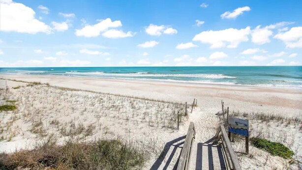 Ein Strand mit einer Holzpromenade, die zum Ufer führt, Sanddünen und einem klaren, blauen Himmel.